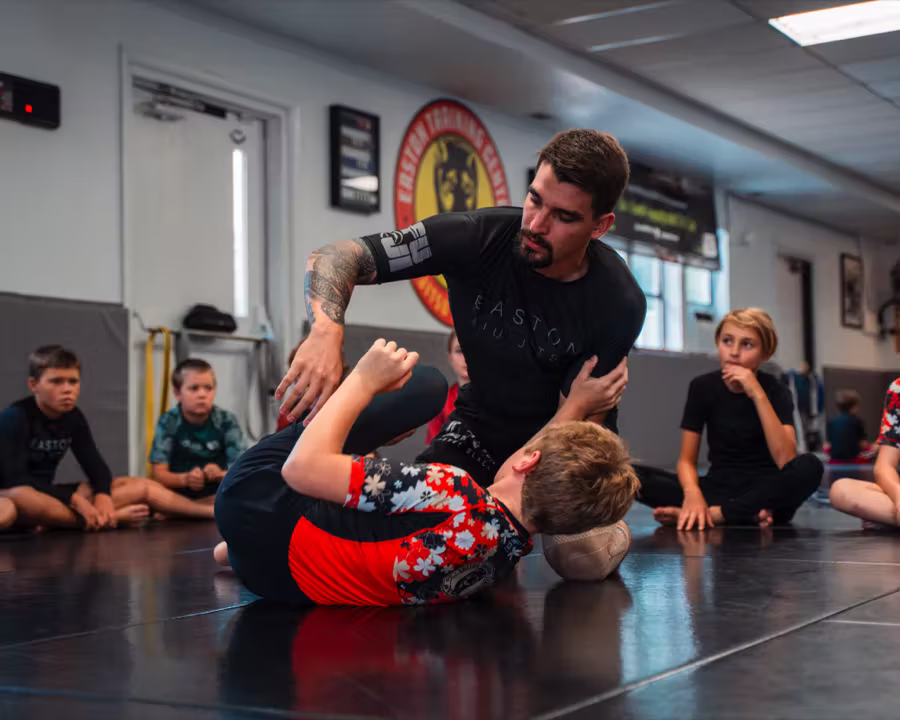 A coach demonstrating a Jiu Jitsu technique on a young student while the rest of the kids class watches.