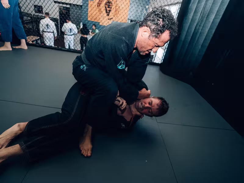 Amal Easton in a black gi, on top of a training partner in a black gi and gripping his collar on the mats, with students in white gis in the background