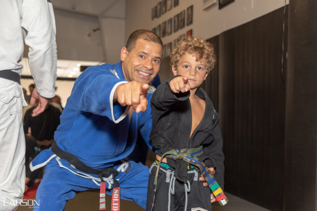 A man and a young boy both wearing Brazilian Jiu Jitsu uniforms point at the camera. Photo by Doug Larson.