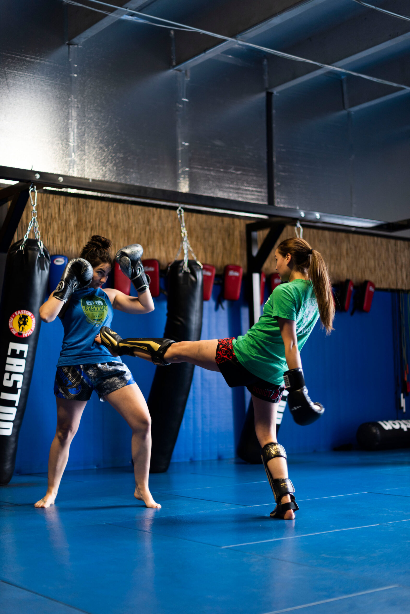 two women wearing boxing gloves face each other, one aims a teep kick at the other's stomach