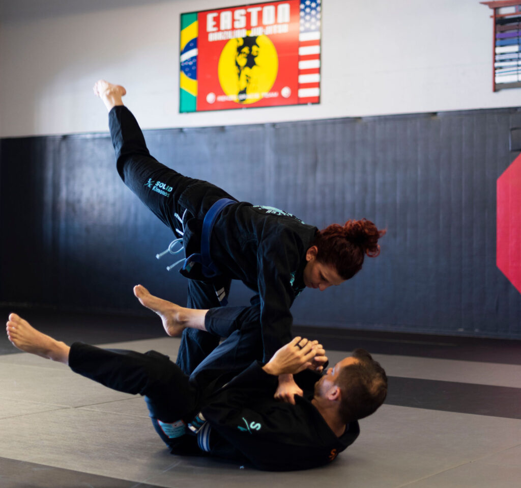 A woman wearing a Brazilian Jiu Jitsu gi hops over a man also wearing a BJJ kimono to pass his guard during Jiu Jitsu class at Easton Training Center Longmont.
