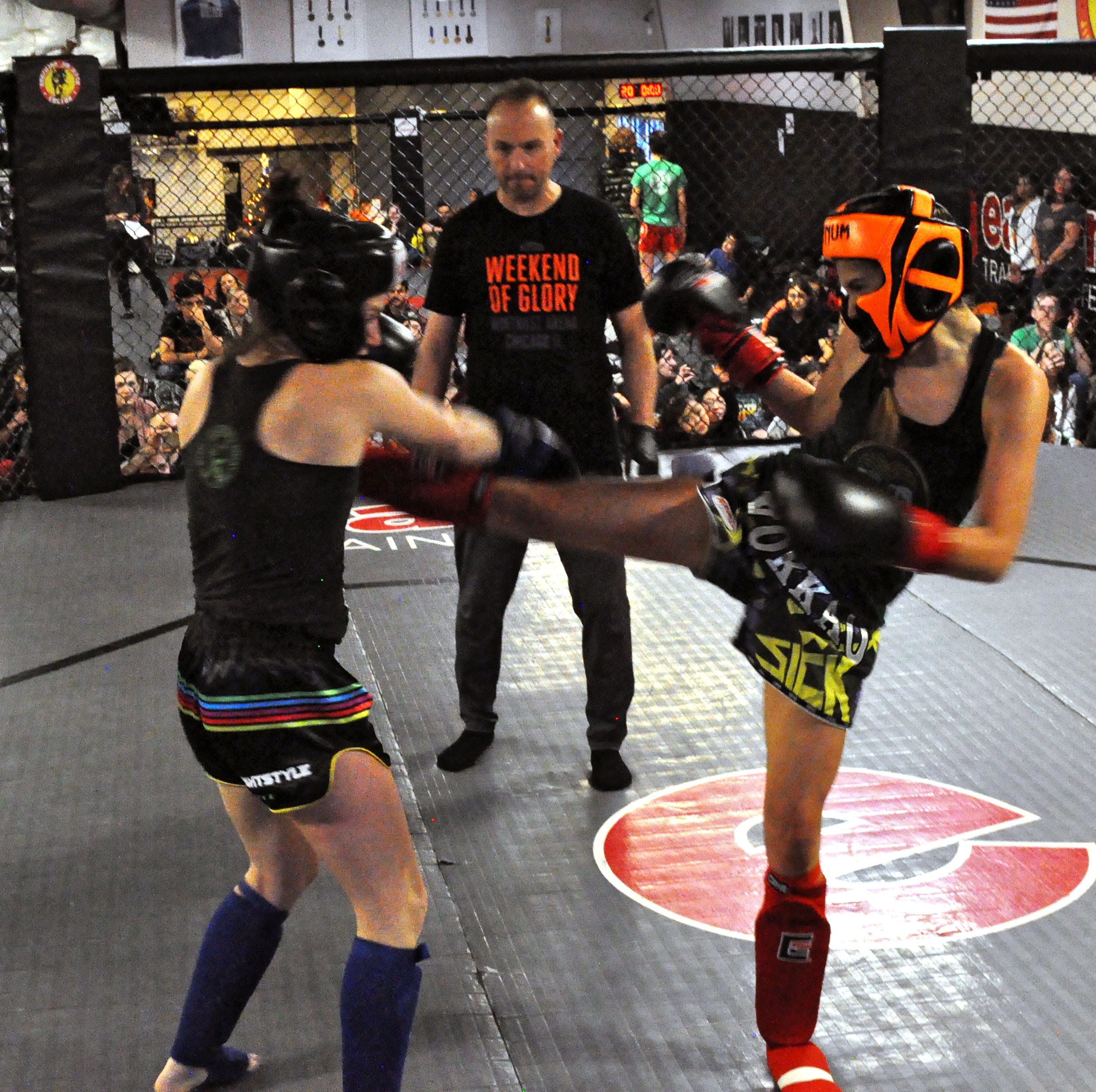 Two women wearing protective gear in the Easton Muay Thai smoker. One kicks the other while a coach looks on.