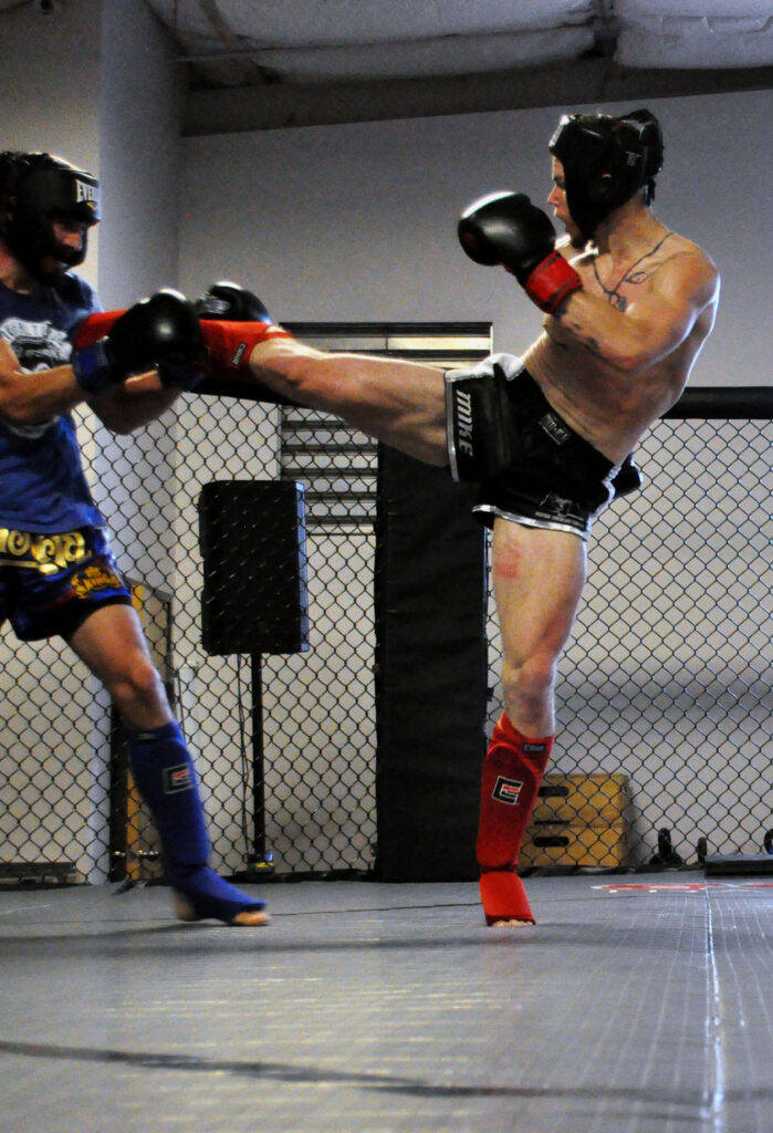 A man wearing Thai shorts, gloves, and protective gear kicks another man in their fight at the Easton Muay Thai smoker.