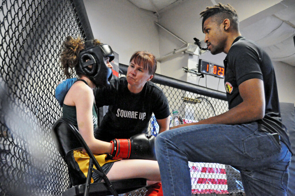 A woman wearing Muay Thai gloves and headgear sits on a chair talking with her coaches between rounds of her Muay Thai smoker fight