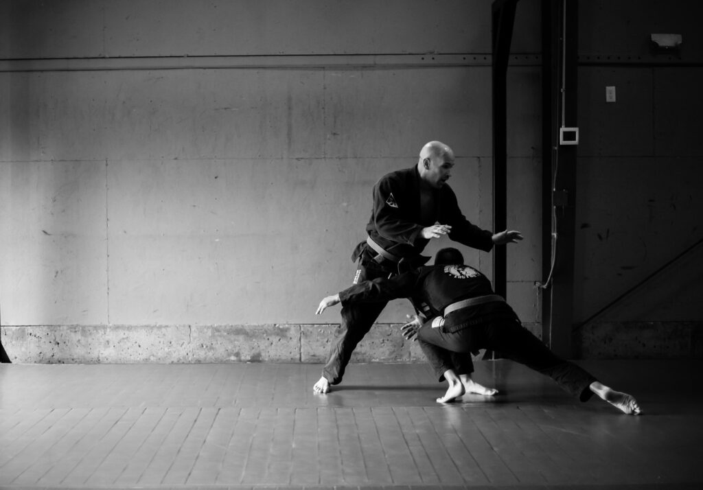 a man sets up a double-leg takedown on another man. Both are wearing Brazilian Jiu Jitsu uniforms.