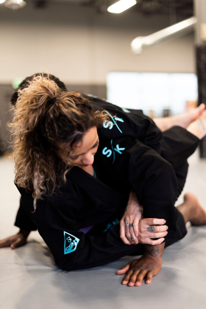 A woman wearing a brazilian jiu jitsu gi sets up a kimura shoulder lock.