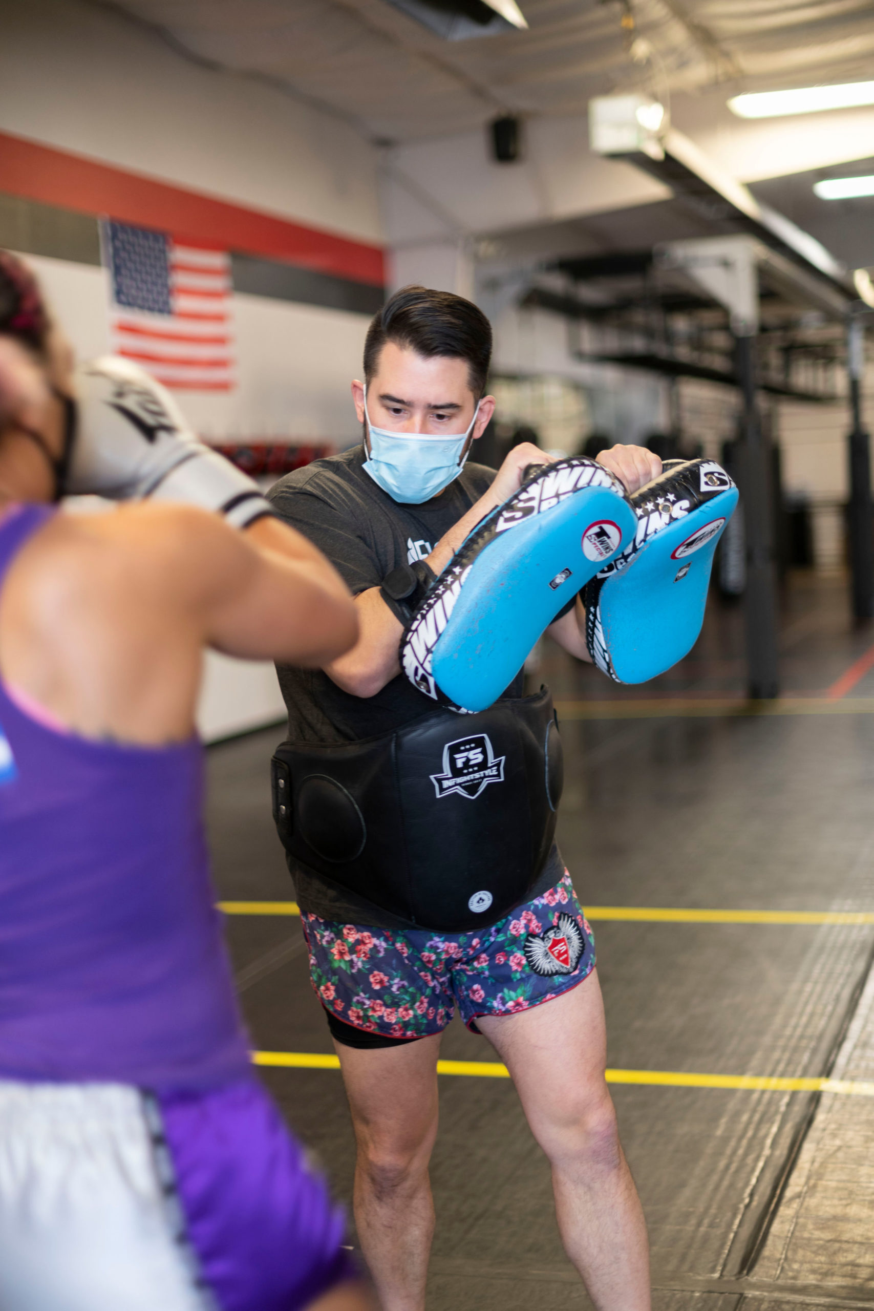 A man wearing a surgical mask holds Thai pads as a woman prepares to kick them