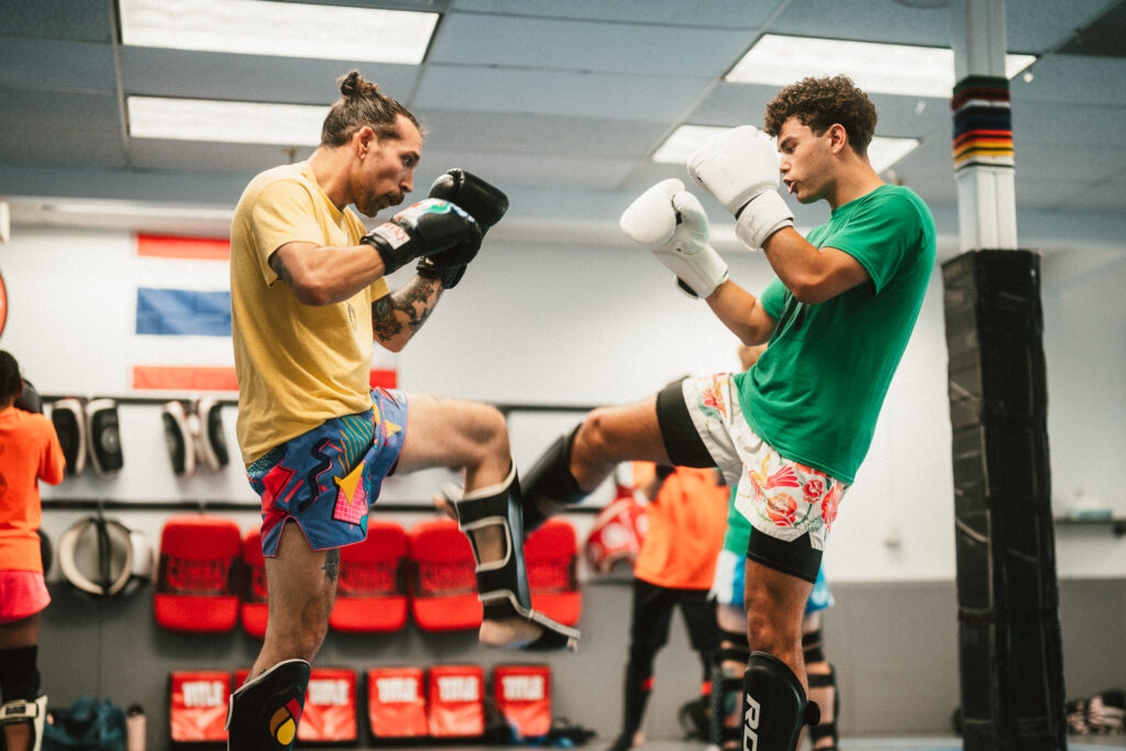 two men in muay thai gloves and shin guards. one checks the other's leg kick.