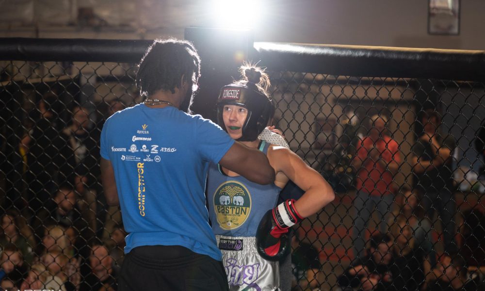Muay Thai coach stands facing his student who is wearing head gear and boxing gloves in the ring between rounds of a Muay Thai fight