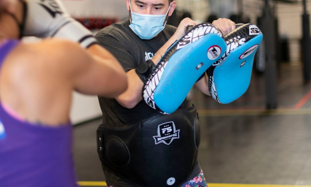 Muay Thai mask Sean kick thai pads A man wearing a surgical mask holds Thai pads as a woman prepares to kick them