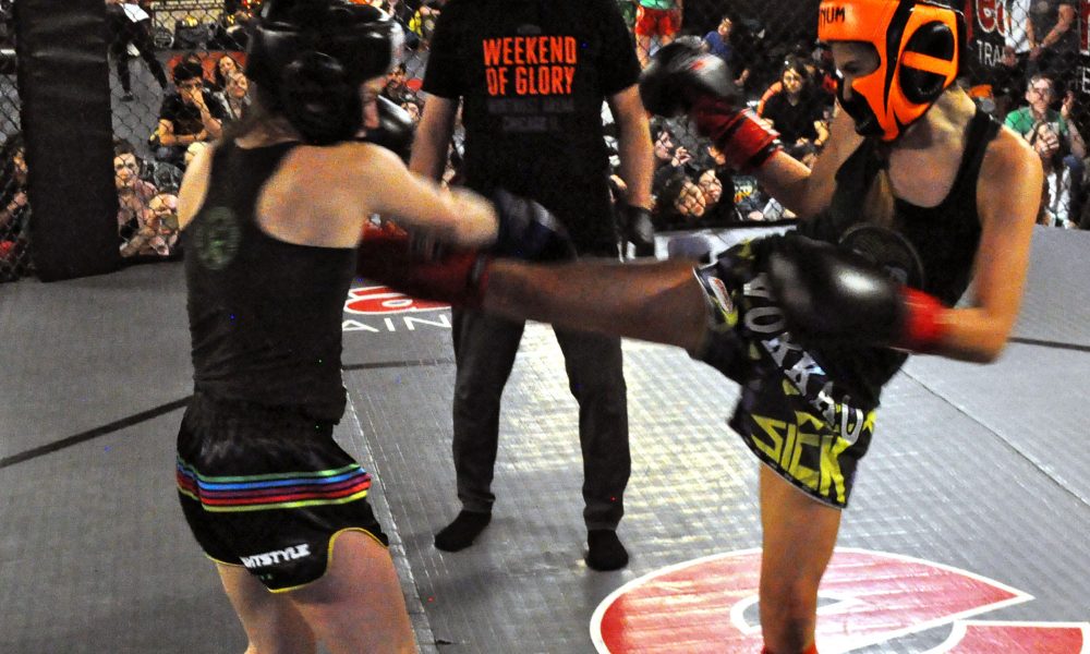 Two women wearing protective gear in the Easton Muay Thai smoker. One kicks the other while a coach looks on.